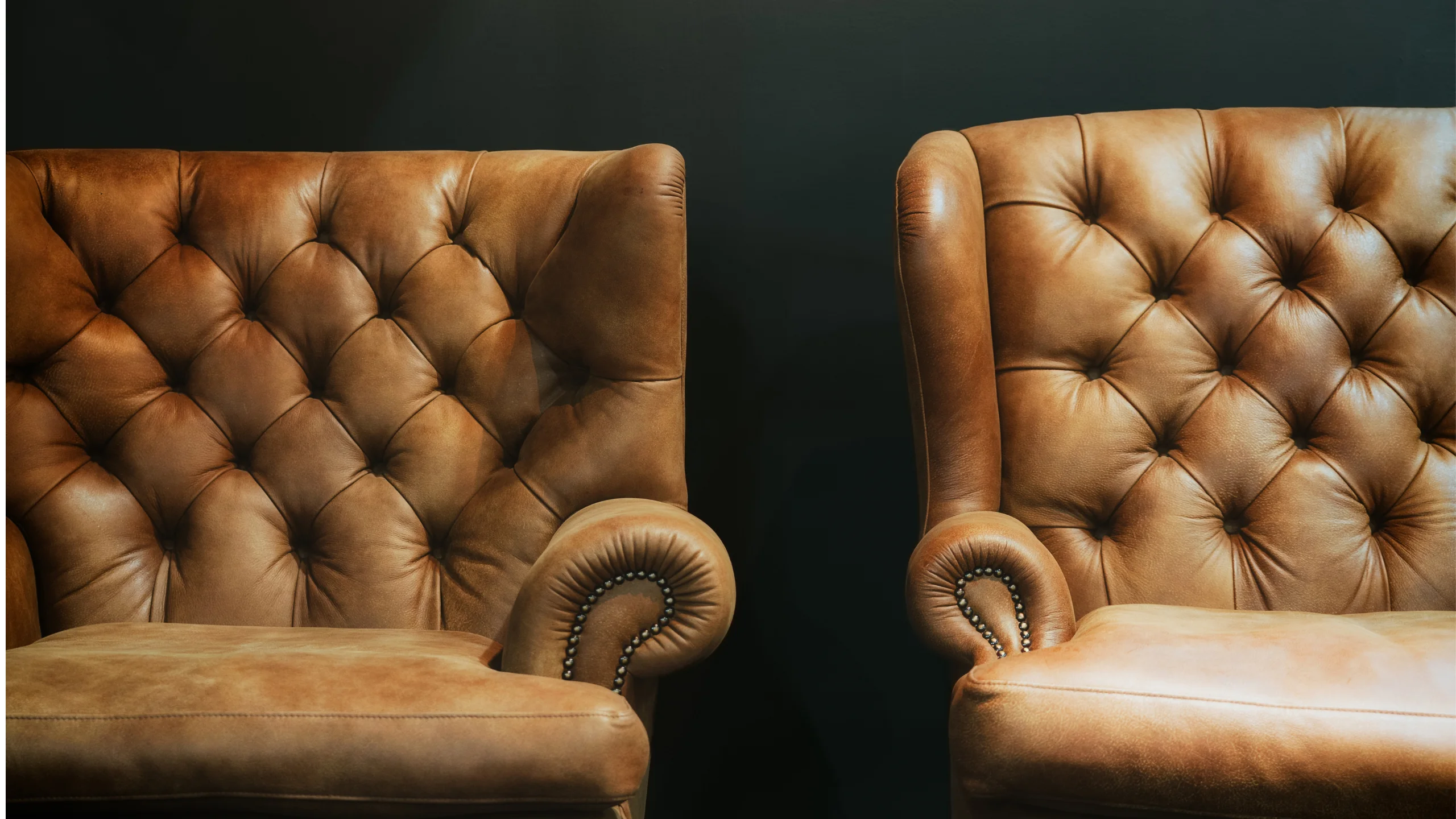 Two tan tufted leather wingback chairs against a dark wall