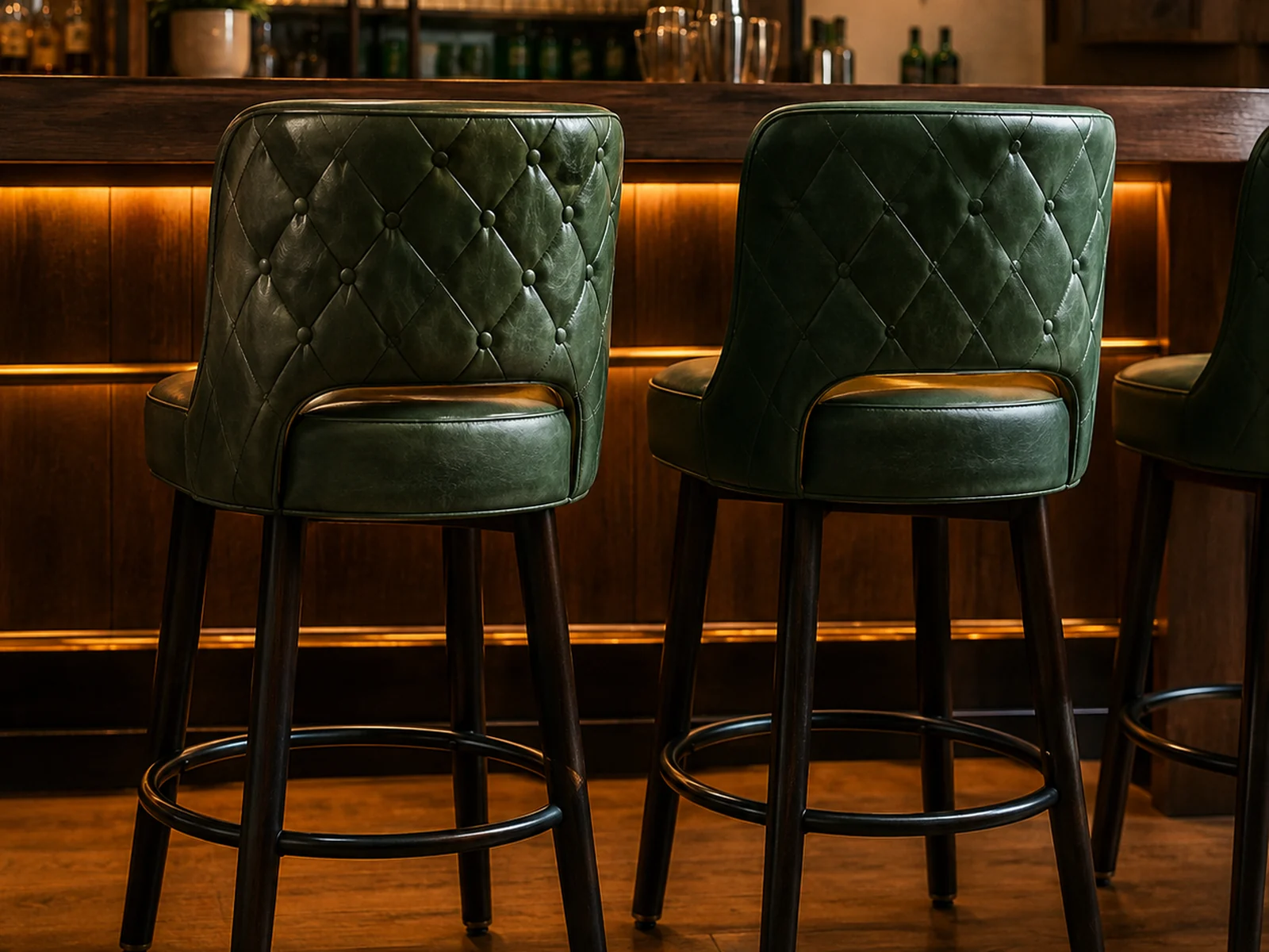 Two green diamond-quilted leather bar stools at a timber bar with a brass footrail