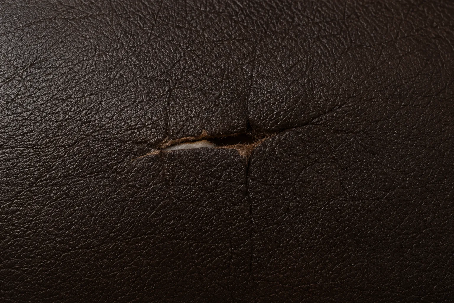 A small puncture torn through a dark brown leather seat, edges lifted around it