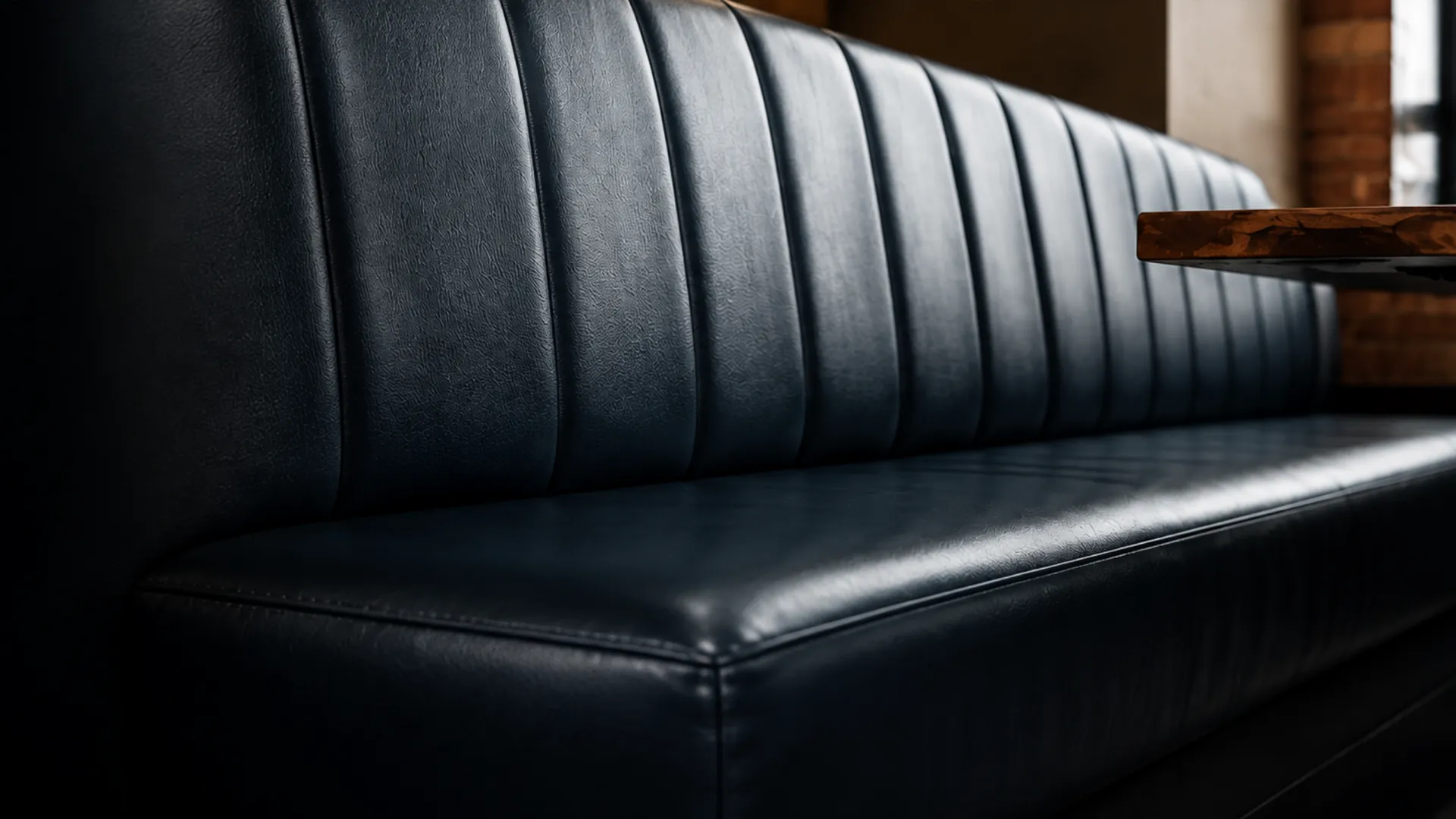 Channel-tufted navy vinyl banquette running the length of a restaurant wall, the vertical flutes catching an even sheen and a walnut table edge crossing the frame