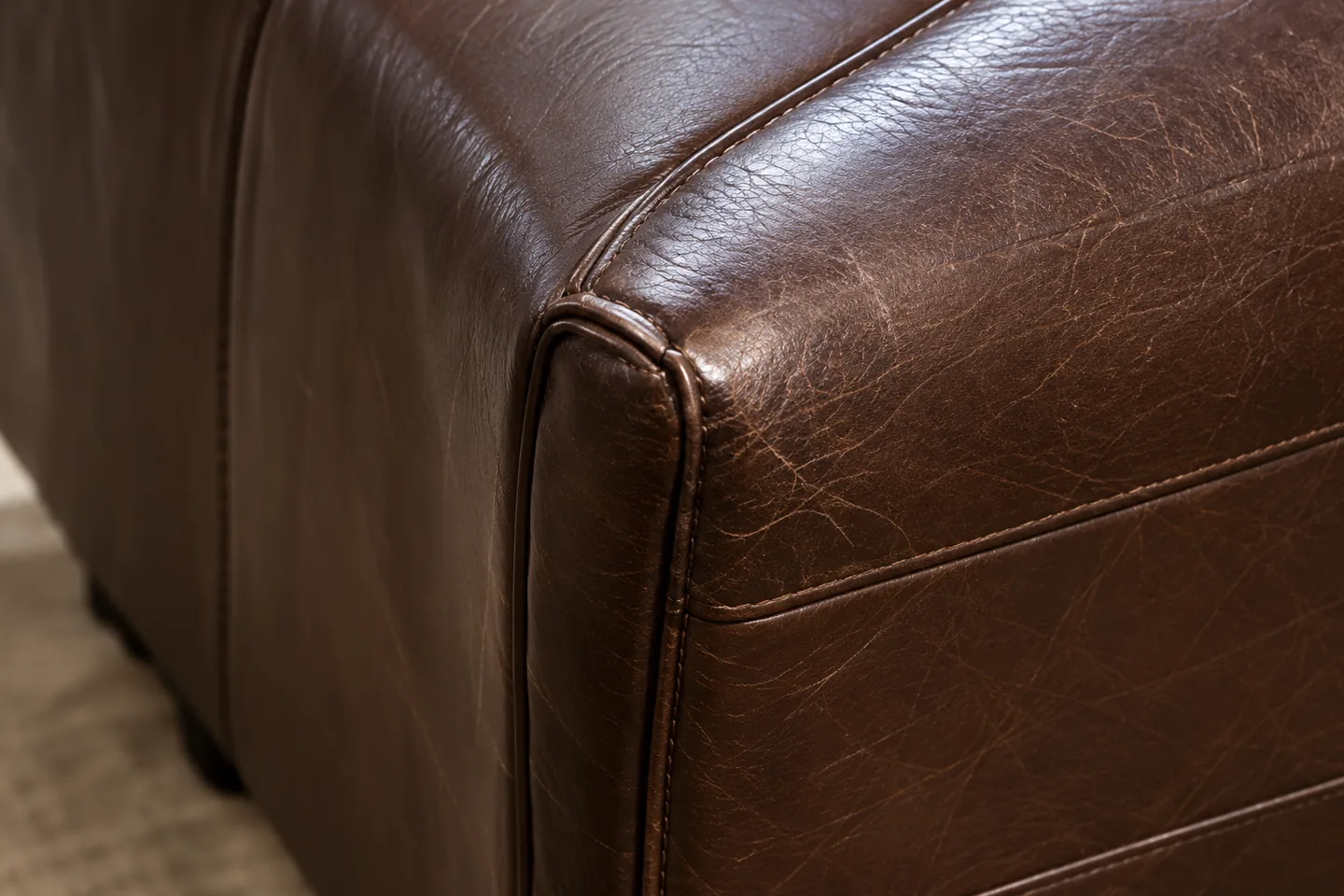 Corner of a brown leather sofa where the outside arm meets the outside back, the surface finely crazed along a welted seam