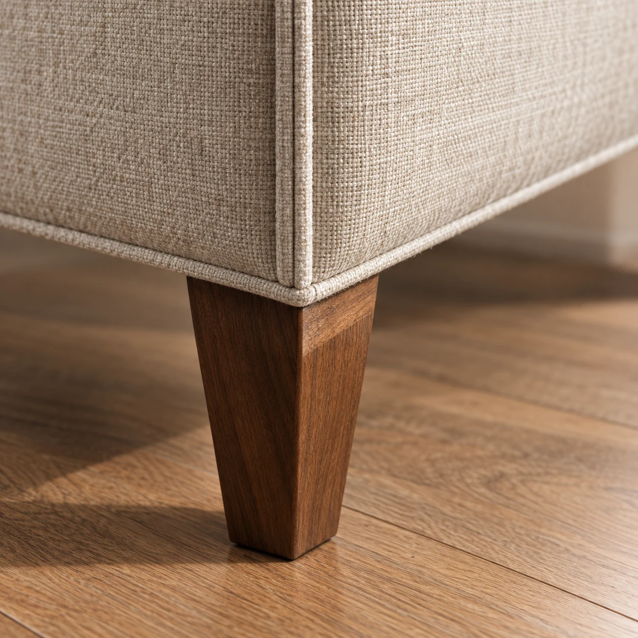 Tapered walnut chair leg under a welted linen corner on a wood floor